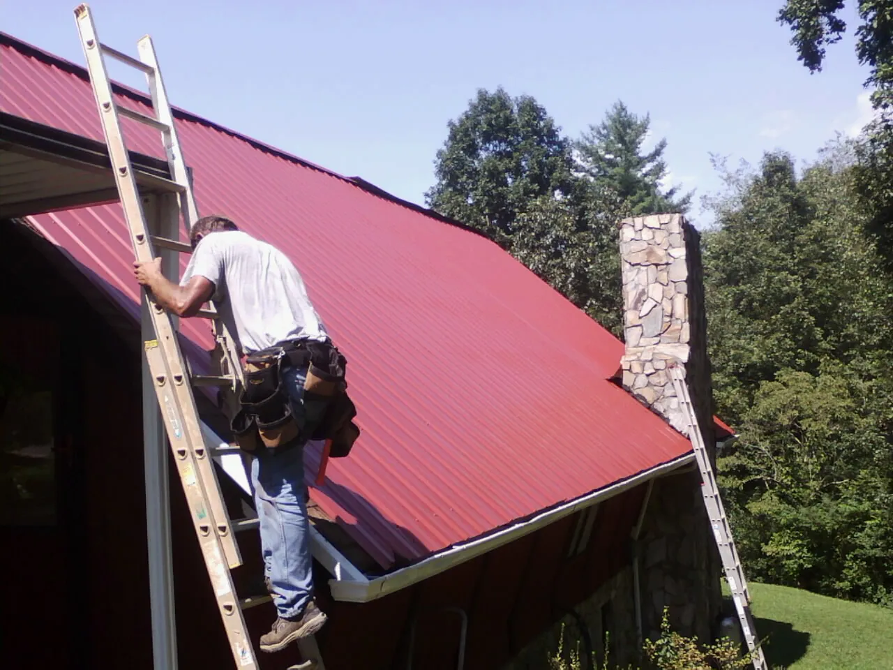 Roofer on a ladder beside a red metal roof and stone chimney — Soffit & Fascia Repair by Roofing Brothers in Pierre Part